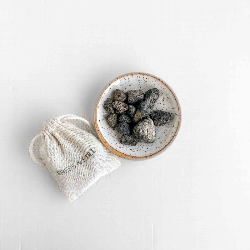Flat lay of small lava stones in a speckled glaze pottery dish, alongside a muslin cotton bag (with Press & Still stamp) containing another two ounces of lava stones.