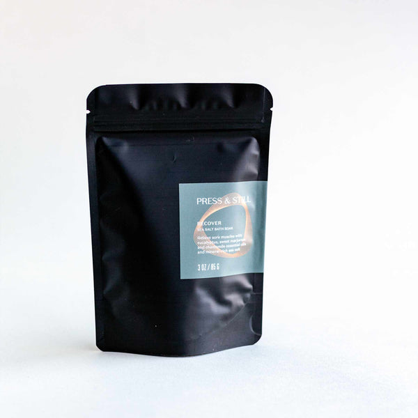 Recover bath soak made with sea salt and essential oils of sweet marjoram, chamomile and eucalyptus. Shown in its matte black foil pouch packaging.