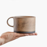 A person holding a Wabi-Sabi stoneware mug with matte glaze in beige with dark bottom.