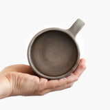 A person holding a Wabi-Sabi stoneware mug with matte glaze in grey-blue. The view is of the top, looking at the inside of the mug. 