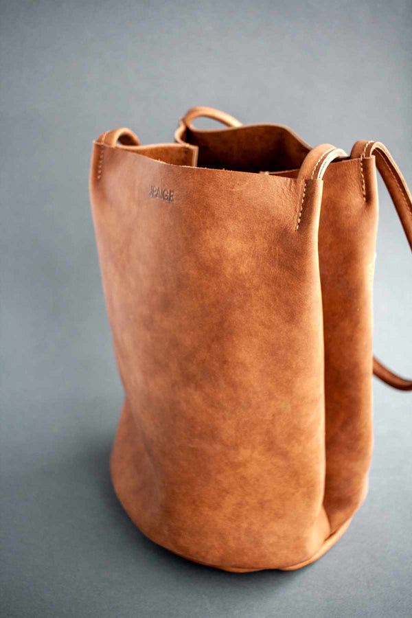 The Fae bucket tote by J Paige & Co. Paige handmade this cocoa-colored bag using leather sourced in the U.S. Shown against a dark grey background.
