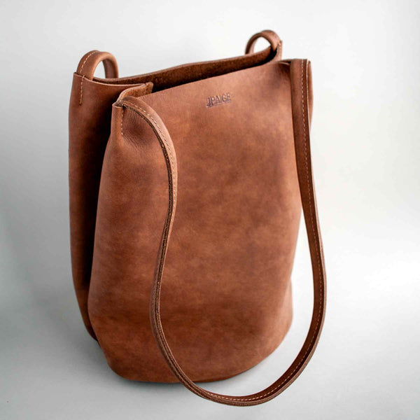 The Fae bucket tote by J Paige & Co. This one's cocoa-colored leather (unlined), shown against a light grey background.