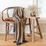 The recycled wool knee blanket in coffee draped over a vintage chair, next to a rustic stool.