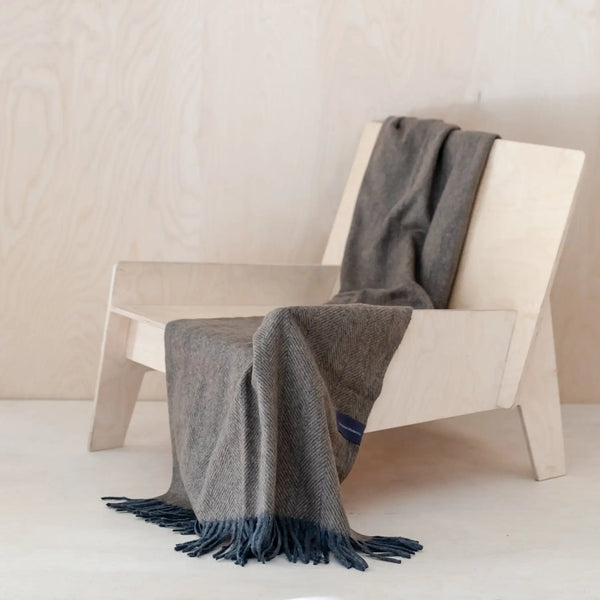 The lovely coffee-colored herringbone knee blanket made of recycled wool is draped over a modern plywood chair. The charcoal - almost blue - fringe is what makes this blanket so unique.