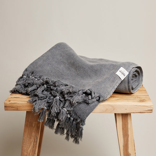 Charcoal vintage wash 100% Turkish cotton throw with herringbone pattern and fringed tassels on wooden stool.